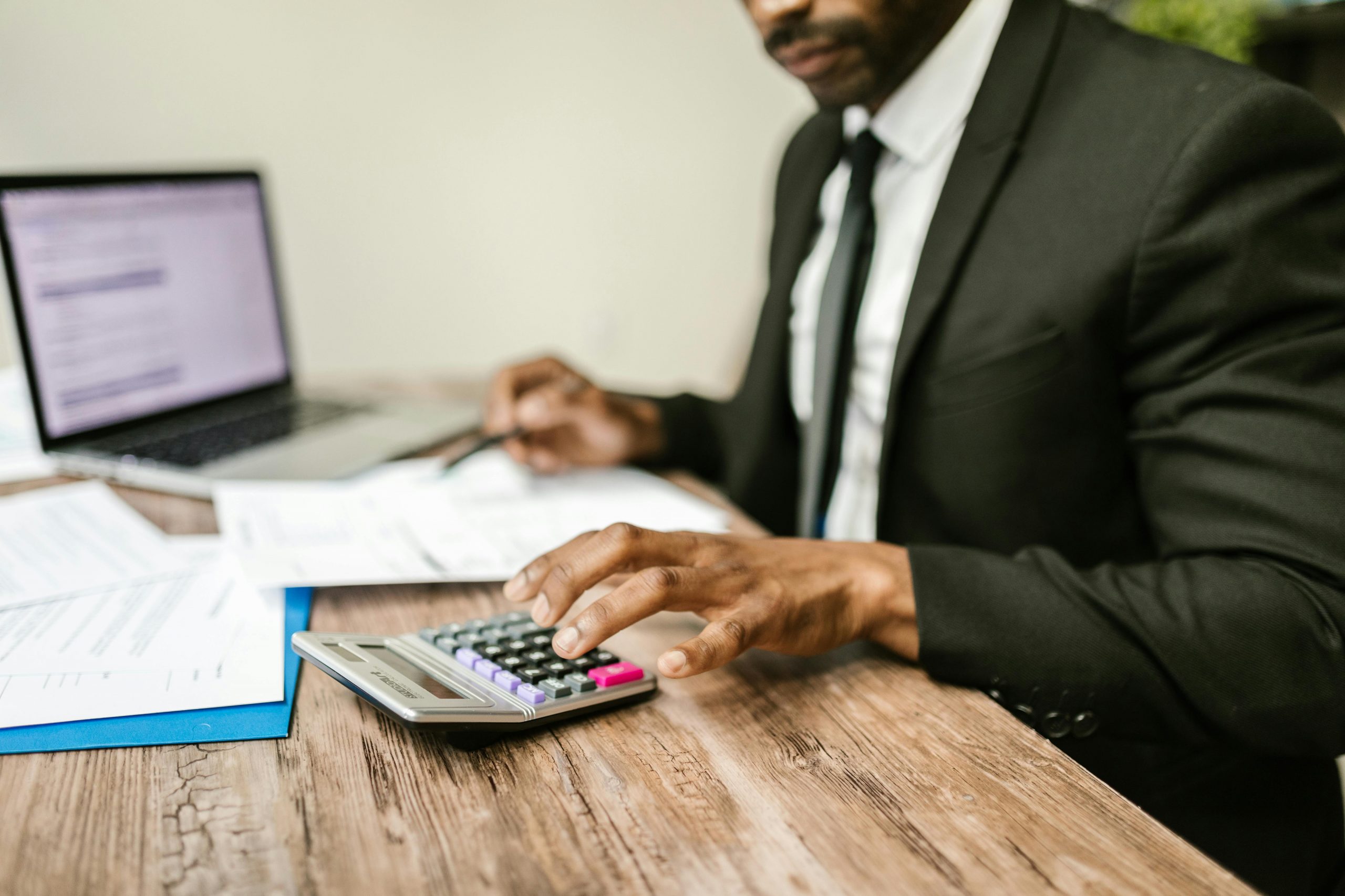 man in a suit working at a desk with a laptop, papers, and a calculator